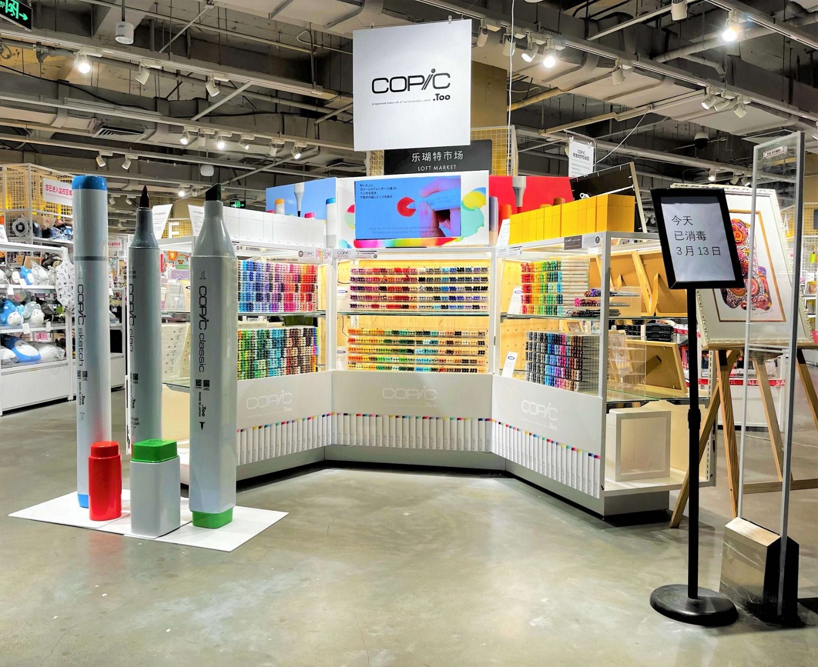 A COPIC stand in a Loft store: walls of coloured markers, three oversized markers on the floor and a framed illustration on an easel.