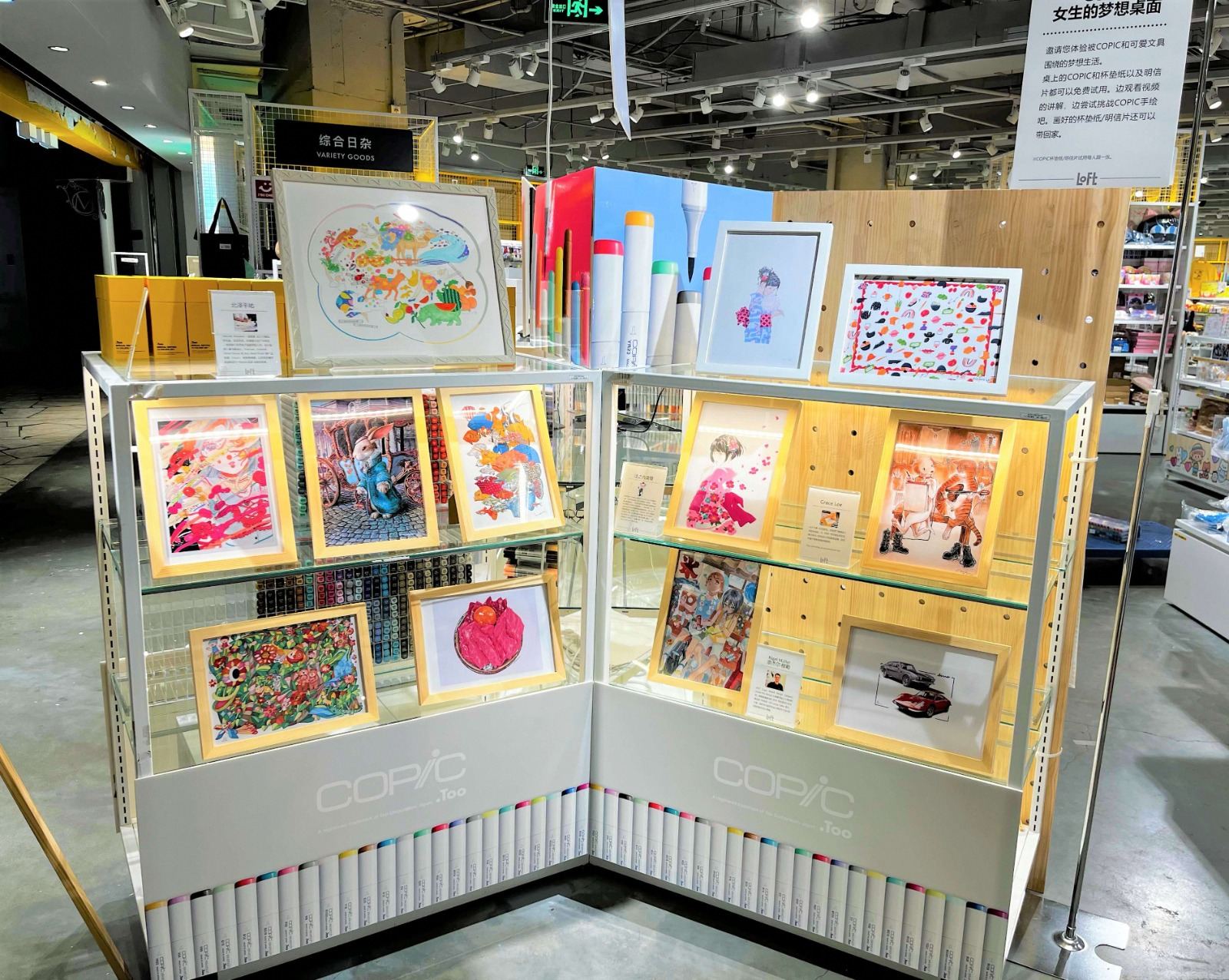 Two glass display cases holding about a dozen framed illustrations, topped with COPIC marker displays, in a Loft store.