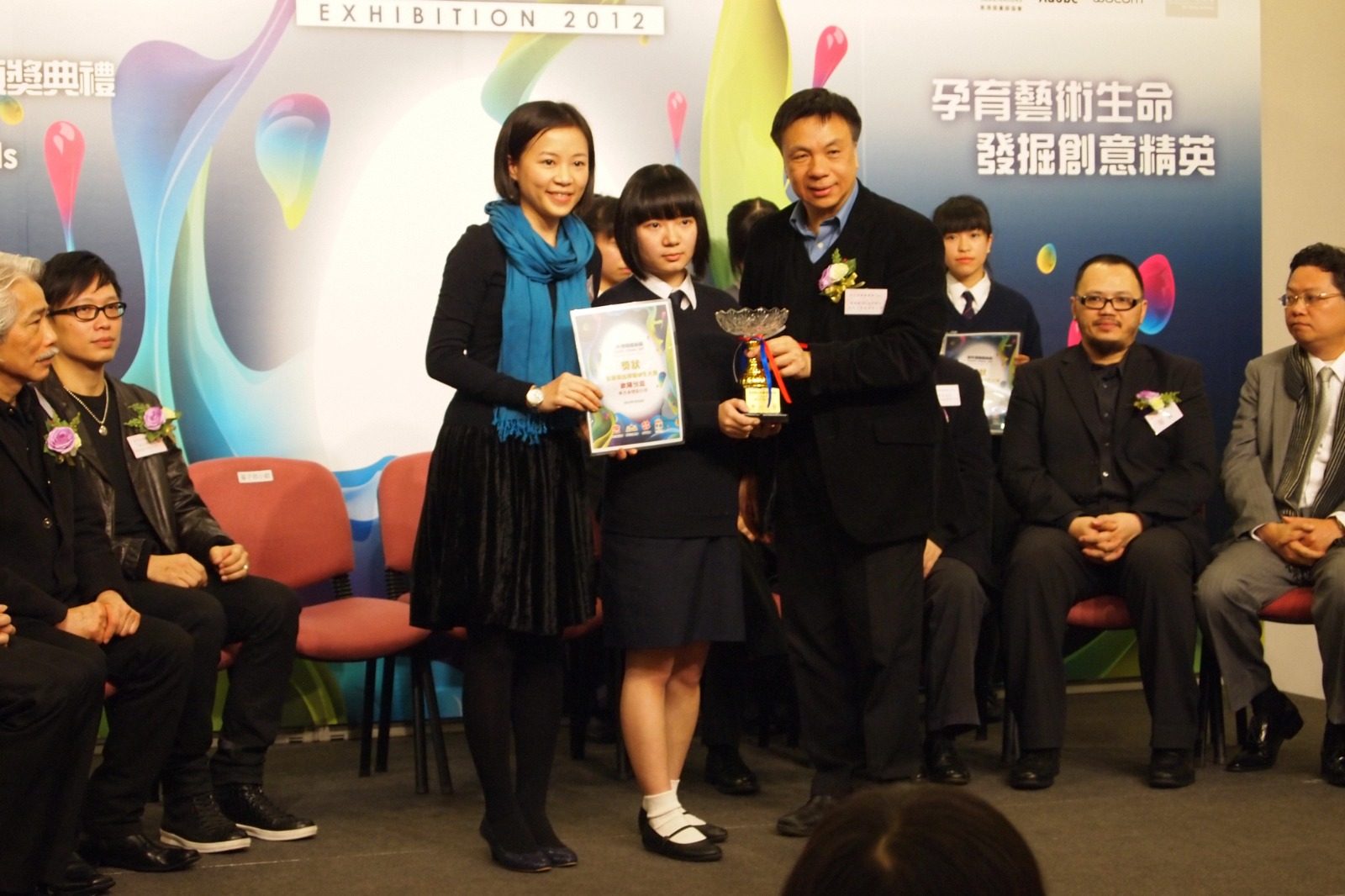 A student in school uniform receives a certificate and a trophy from two organisers on a stage, in front of a colourful board reading Exhibition 2012.
