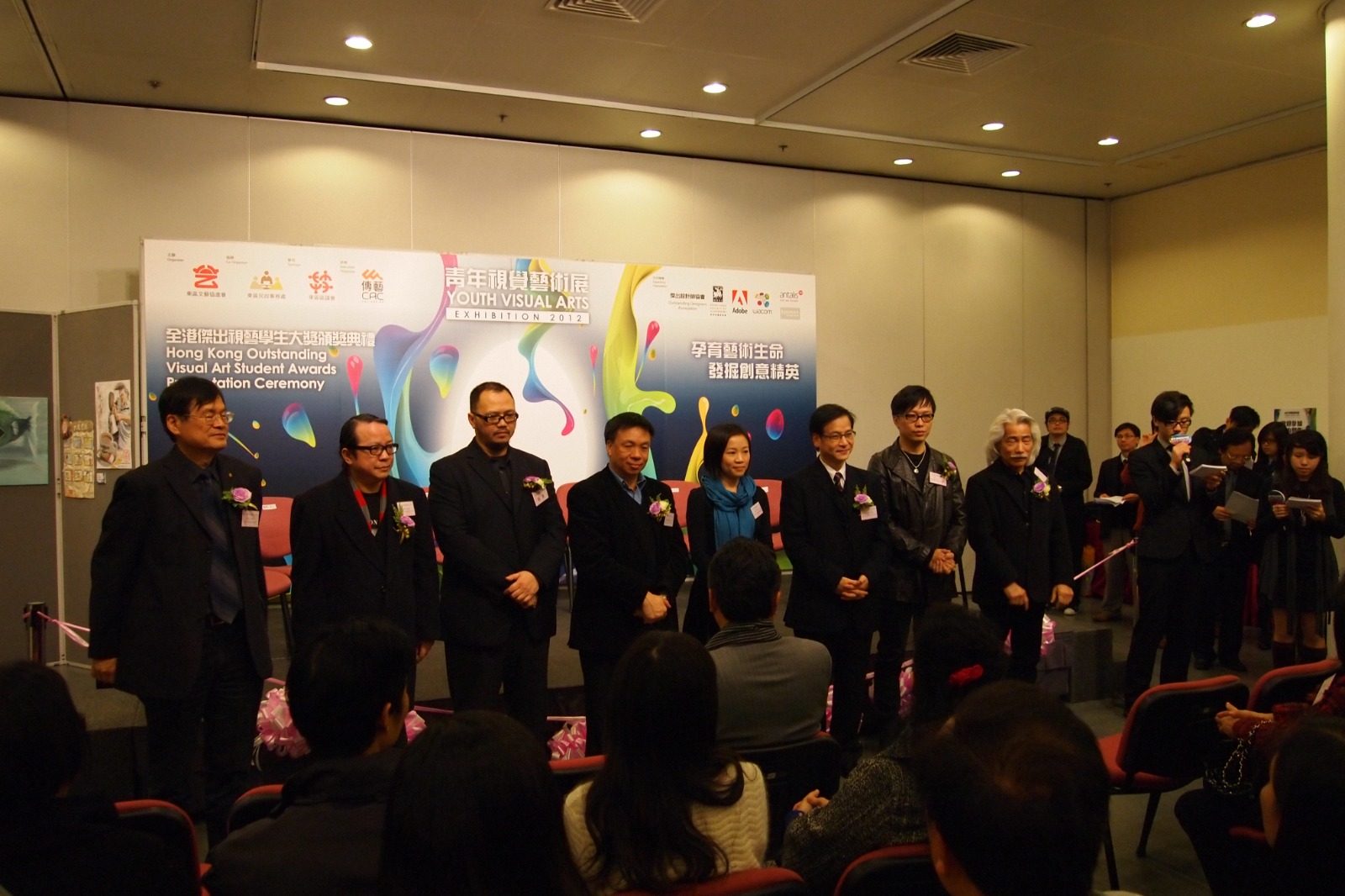 Eight panel members and organisers lined up on a stage, in front of the bilingual board reading Hong Kong Outstanding Visual Art Student Awards Presentation Ceremony, Youth Visual Arts Exhibition 2012.